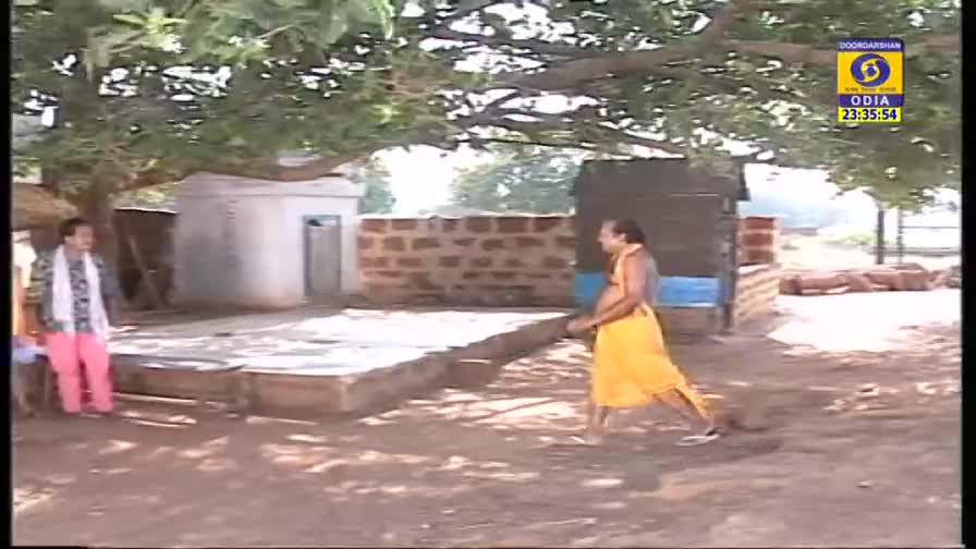 A man in a yellow garment strides across the dirt, his bare feet slapping the ground. Another man, wearing pink pants and a white scarf, stands still, watching.
