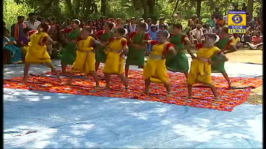 On a colorful rug, dancers in yellow and green costumes are performing, their arms and legs moving in unison. The DD Odia logo is visible, and a crowd watches from the background.
