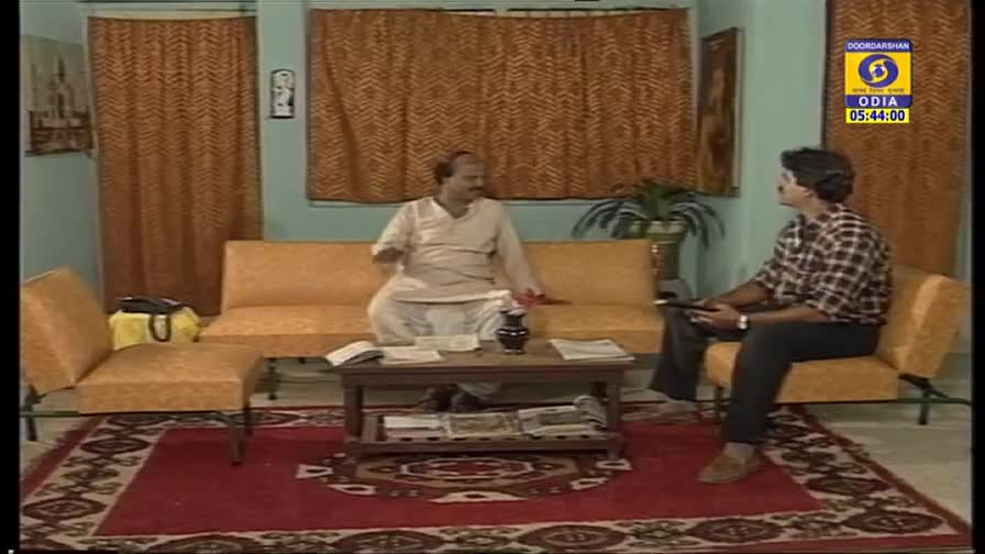 Two men sit in what looks like a living room, one in a white kurta gesturing while the other holds a remote. The DD Odia logo in the corner confirms this is a broadcast from India.
