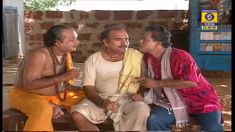 Three men sit on a bench, engaged in what looks like a heated discussion; the man on the right gestures emphatically. The scene is broadcast on DD Odia.
