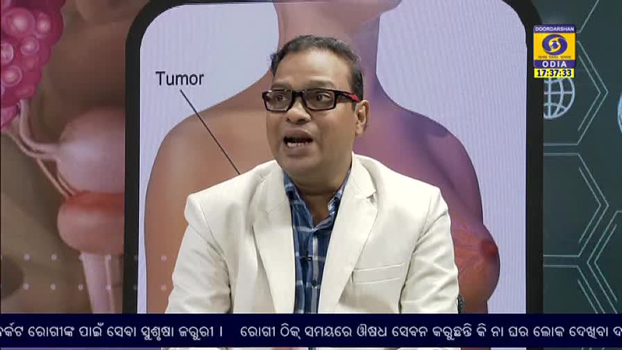 A man in glasses, wearing a blazer, speaks animatedly on DD Odia. Behind him, a diagram of a body highlights a tumor.
