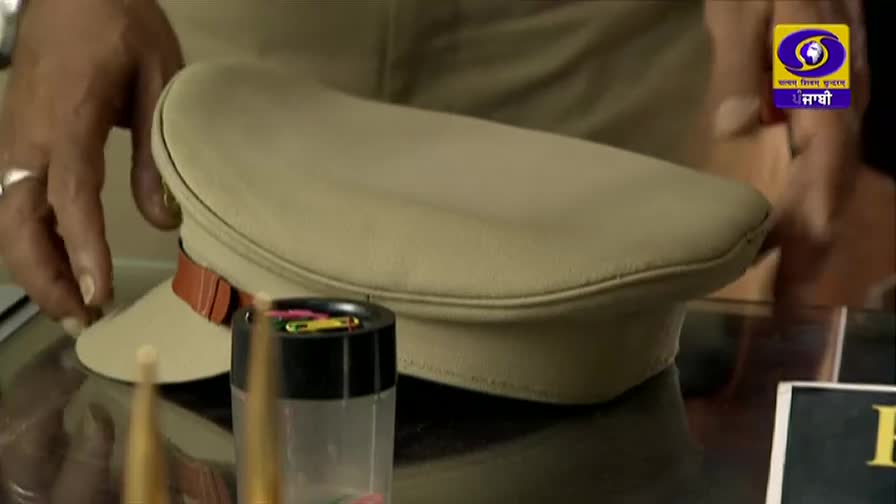 A police cap rests on a desk, its brim angled towards a small container of colorful paperclips. The DD Punjabi logo is visible in the corner, and a hand is about to touch the cap.
