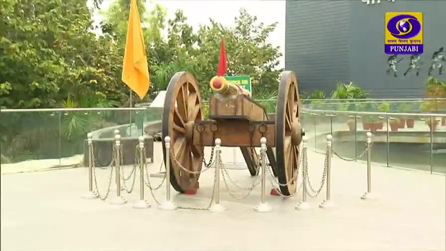 A replica cannon sits on display, framed by a chain barrier and a glass wall. The DD Punjabi logo is visible in the corner, and flags wave gently in the background.
