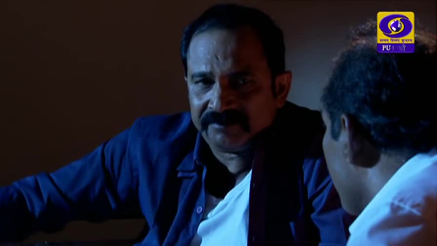 A man in a blue shirt leans forward, his gaze intense as he speaks to someone out of frame. The DD Punjabi logo is visible in the corner, suggesting this is a scene from a television drama.
