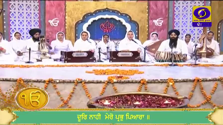 Musicians in white robes and turbans are playing instruments and singing on a stage decorated with flowers and candles. The scene is being broadcast on DD Punjabi, a channel from India.
