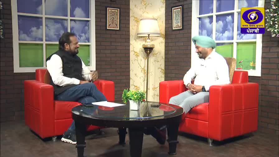Two men are seated in red armchairs, facing each other, engaged in conversation. The set, decorated with a brick-patterned wall and a logo for DD Punjabi, suggests a television interview is underway.
