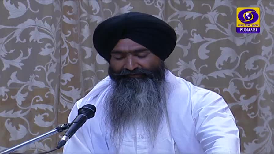 A man in a black turban and white robe sits at a microphone, his eyes closed and head bowed. The DD Punjabi logo of India is visible in the corner, suggesting a broadcast.
