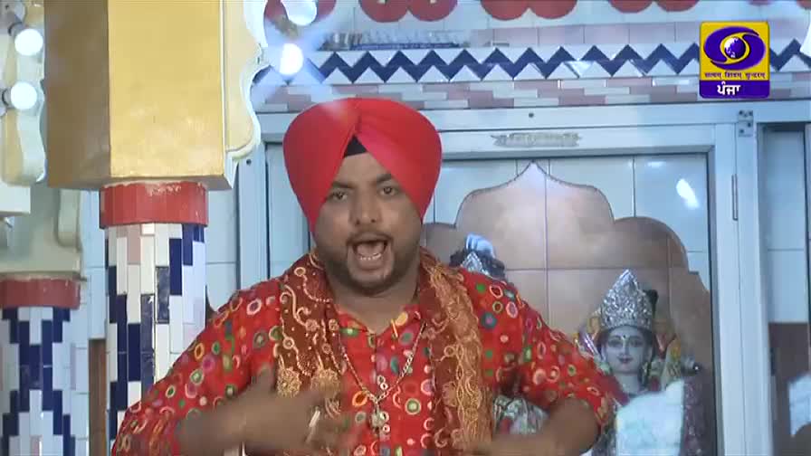 A man in a red turban and patterned shirt gestures emphatically, his mouth open as if singing. The DD Punjabi logo sits in the corner, and a statue of a deity is visible behind him.
