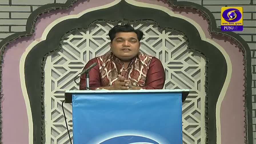 A man in a decorative vestment stands behind a blue podium, speaking into a microphone. Behind him, a patterned wall and the DD Punjabi logo indicate a broadcast from India.
