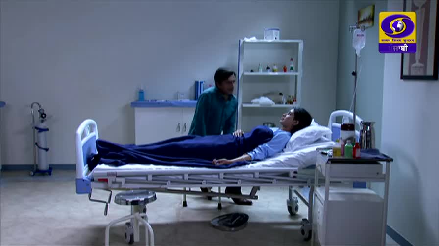 In a hospital room, a woman lies in a bed, covered by a dark blue blanket, while a man in a teal shirt stands beside her. The DD Punjabi logo is visible, indicating this is likely a scene from an Indian television drama.
