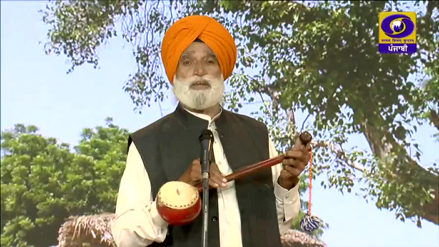 A man in a bright orange turban stands before a microphone, holding a stringed instrument. He appears to be on a DD Punjabi set, with a backdrop of a tree and sky.
