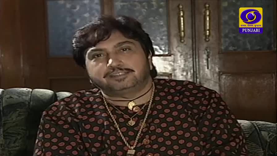 He's sitting on a patterned sofa, facing the camera, wearing a polka-dotted shirt and multiple gold chains. The DD Punjabi logo is visible in the corner.
