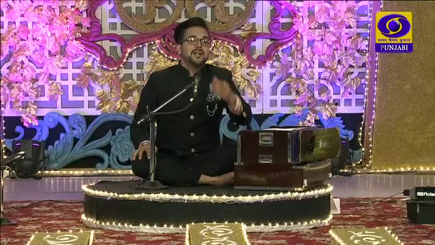 The man, seated on a low platform, gestures with his right hand as he speaks. Behind him, the set is elaborately decorated with ornate floral and geometric designs, lit by soft, colored lights.
