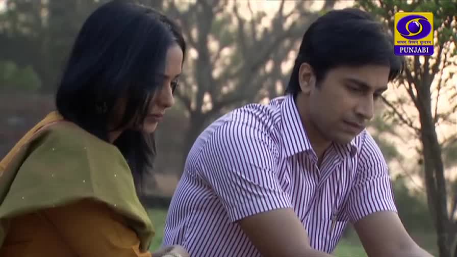 The woman is turned away, her dark hair framing her face as she looks down. The man in the striped shirt sits beside her, his gaze also fixed downwards, the DD Punjabi logo visible in the corner.
