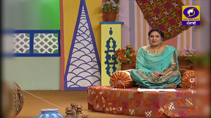 A woman in a teal salwar kameez sits cross-legged on a patterned cushion, smiling at the camera. The DD Punjabi logo is visible in the corner, indicating a television studio in India.
