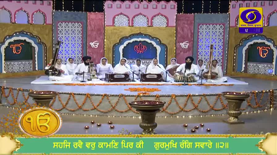 On a stage decorated with lights and floral garlands, a group of musicians in white robes and turbans play instruments. The DD Punjabi logo is visible in the corner, and the broadcast is underway.
