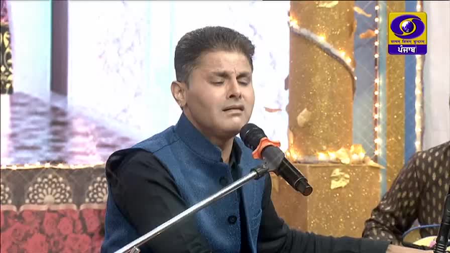 A man in a blue vest sings into a microphone, eyes closed, on a DD Punjabi program. The background is ornate and golden, with the channel's logo visible in the corner.
