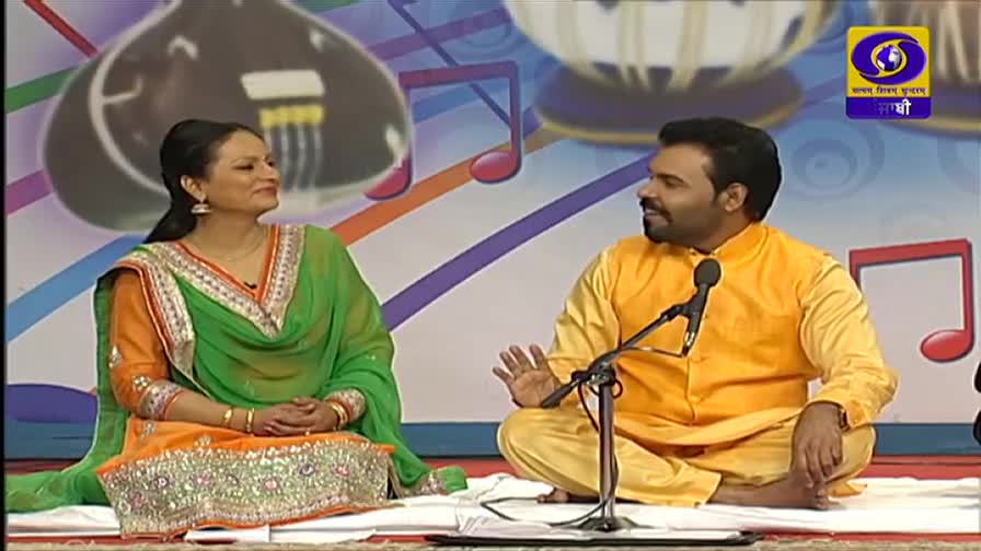 The man in the yellow kurta gestures with his hands as he speaks into the microphone. Seated beside him, the woman in the green and orange salwar kameez smiles, listening intently.
