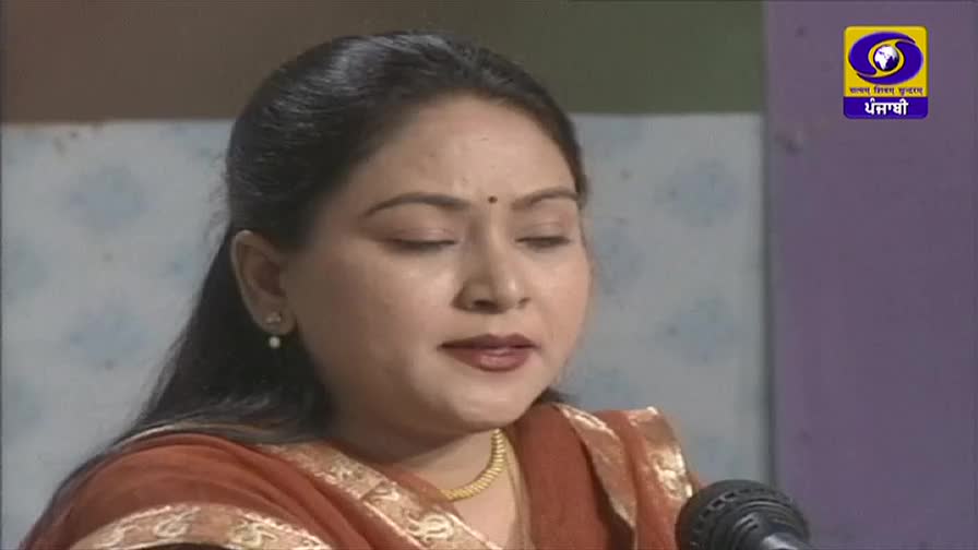 A woman in a rust-colored shawl is speaking into a microphone, her eyes closed. The DD Punjabi logo is visible in the corner, suggesting a broadcast from India.
