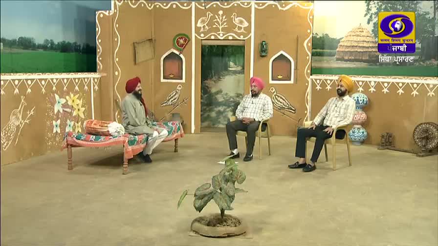 Three men in turbans are seated on a set decorated to look like a rural Indian home, presumably for a DD Punjabi program. The man on the left sits on a cot, while the other two are in chairs, engaged in a discussion.
