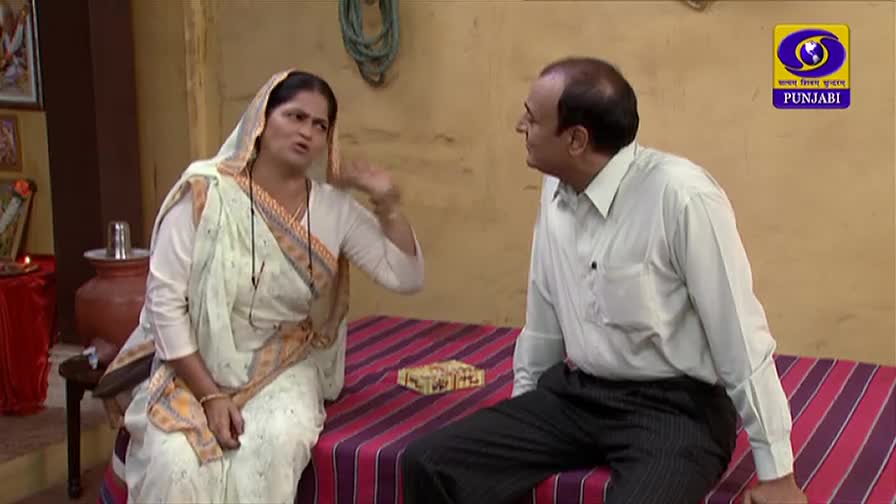 A woman in a white sari gestures emphatically, her hand raised as she speaks to a man beside her. He listens, seated on a patterned bed, as the DD Punjabi logo shines in the corner.
