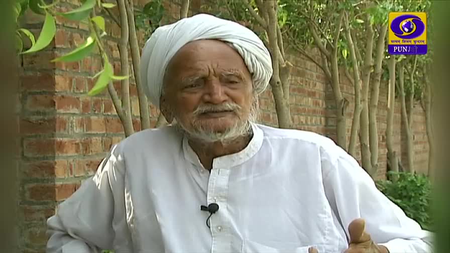 An elderly man in a white turban and shirt gestures with his hands as he speaks. The DD Punjabi logo sits in the corner, and a brick wall and some trees form the backdrop.
