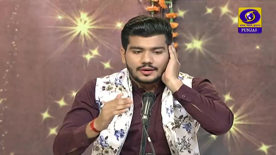 The man, wearing a floral vest over a maroon shirt, gestures with one hand and holds the other to his ear. He's speaking into a microphone, likely on a DD Punjabi program.
