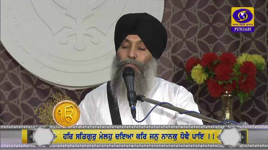 A man in a black turban speaks into a microphone, his eyes closed as he recites, likely on a broadcast from DD Punjabi. The backdrop is a simple, patterned wall, and a vase of flowers sits to the side.
