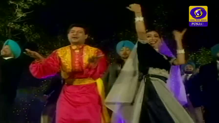 A man in a bright pink and gold outfit gestures with his hands as he sings. A woman in a black and white dress raises her arms, while others in turbans and suits stand behind them.
