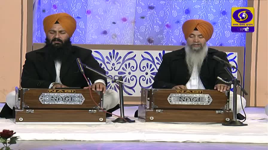 Two men in turbans sit facing each other, their fingers moving over the keys of the harmoniums. The DD Punjabi logo is visible in the corner of the screen.
