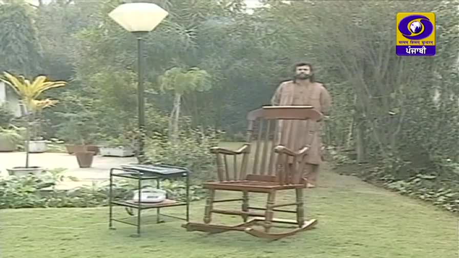 A man in a brown kurta stands behind a wooden rocking chair on a lawn, facing the camera. The DD Punjabi logo is visible in the top right corner, suggesting a television program.
