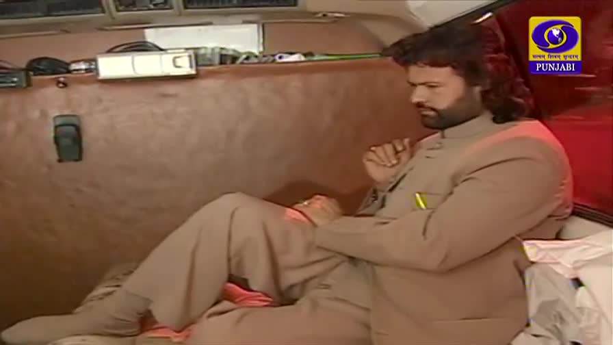 A man in a light suit sits inside a vehicle, looking down with a thoughtful expression. The DD Punjabi logo is visible in the corner, suggesting a scene from India.
