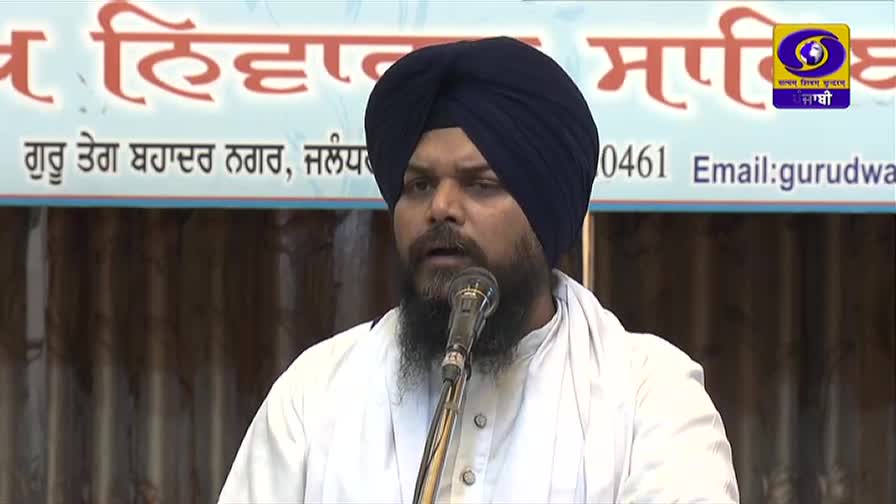 A man with a dark turban and beard speaks into a microphone. The DD Punjabi logo is visible, and a banner in Punjabi is behind him.
