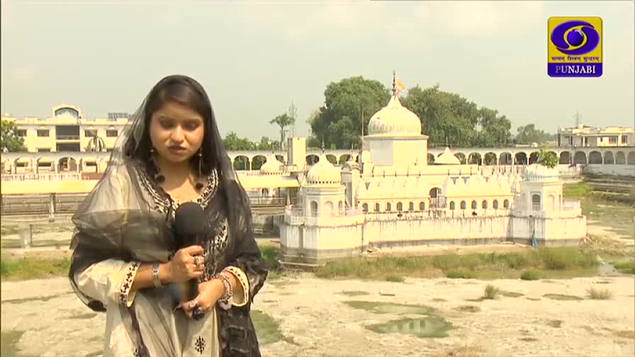 A woman in a scarf and ornate clothing stands with a microphone, reporting in front of a white temple. The DD Punjabi logo sits in the corner, and the scene is set in India.
