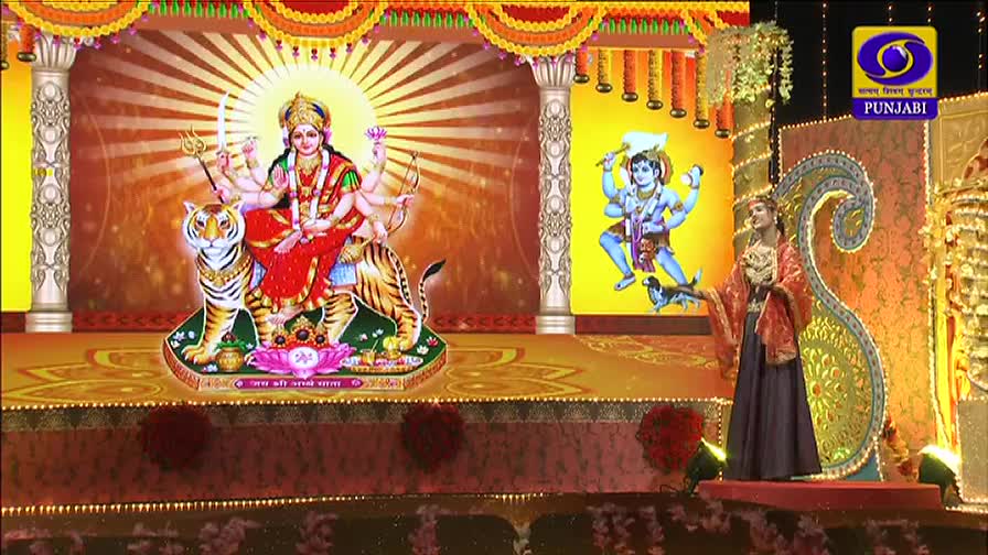 On the stage, a performer in a long dress and ornate jacket gestures while standing before a large, stylized backdrop of Hindu deities. The DD Punjabi logo is visible in the upper right corner.
