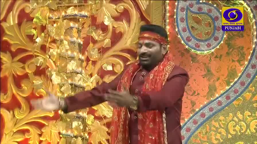 A man in a maroon suit and red scarf gestures with his hands as he speaks. Behind him, a decorative backdrop with gold filigree and the DD Punjabi logo fills the frame.
