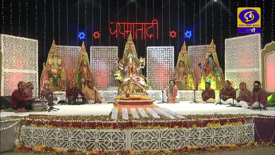 On a stage decorated with lights, musicians in maroon shirts are seated, playing instruments and singing. In the center, a golden statue of a deity sits, flanked by other figures, all under the DD Punjabi logo.
