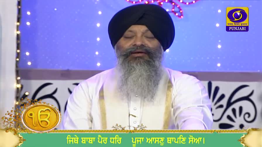 A man in a black turban, eyes closed, is speaking on DD Punjabi. The screen behind him is a soft blue, with a logo in the corner.
