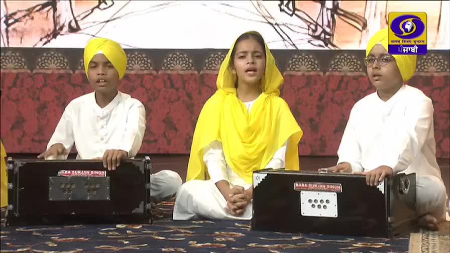 Three children, dressed in white and yellow, sit on the floor, singing. They're playing small harmoniums, their hands moving over the keys.
