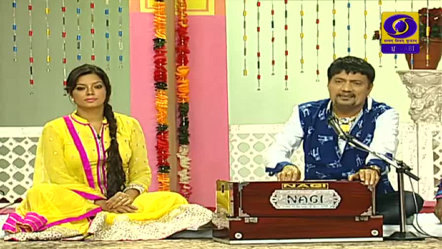 A woman in a yellow outfit sits calmly, while a man plays a harmonium labeled "Nagi." The DD Punjabi logo is visible, suggesting a broadcast in India.
