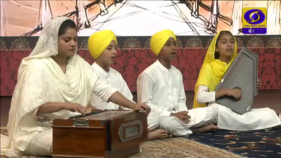 Four young people, dressed in white, sit on the floor, singing. A woman plays a harmonium, and another girl strums a stringed instrument, all on a set for DD Punjabi, the Indian television channel.

