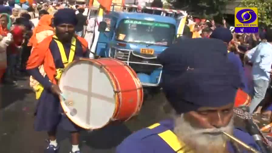 A man in a turban marches forward, striking a large drum with a white face and red rim. Behind him, a blue auto-rickshaw slowly navigates through a crowd, as the DD Punjabi logo appears on the screen.
