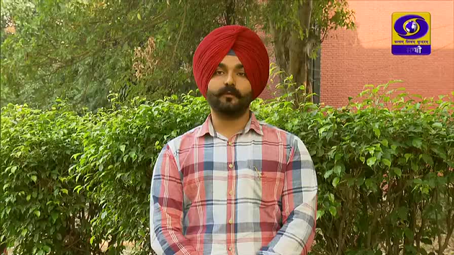 A man in a red turban and plaid shirt stands in front of a green hedge, likely filming for DD Punjabi. The India logo is visible in the upper right corner.
