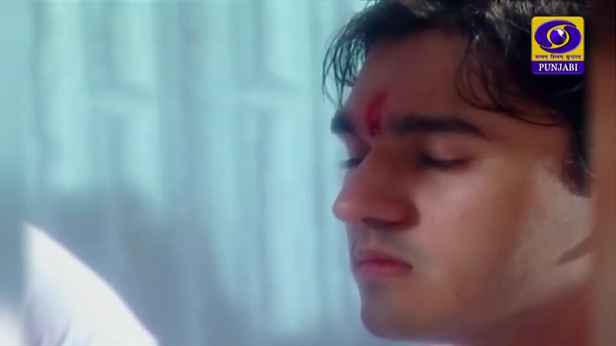 A man with closed eyes and a red mark on his forehead is in focus, likely from a DD Punjabi broadcast. The logo of the channel is visible in the corner.
