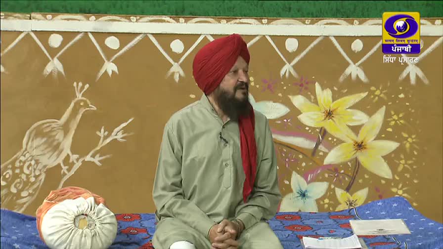 A man in a red turban and beard sits on a patterned cushion, speaking into a microphone. Behind him, a painted backdrop of flowers and a peacock decorates the set of the DD Punjabi program.
