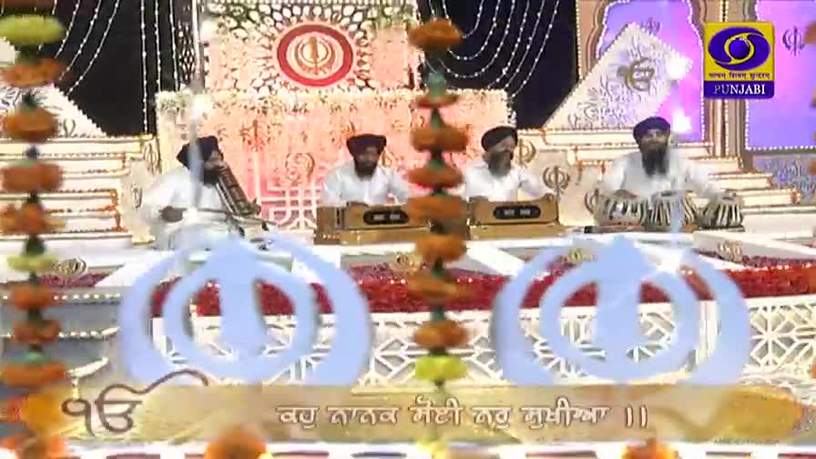 Musicians in white robes and turbans play instruments on a stage, the sounds broadcast on DD Punjabi. A string of orange flowers hangs down in the foreground.
