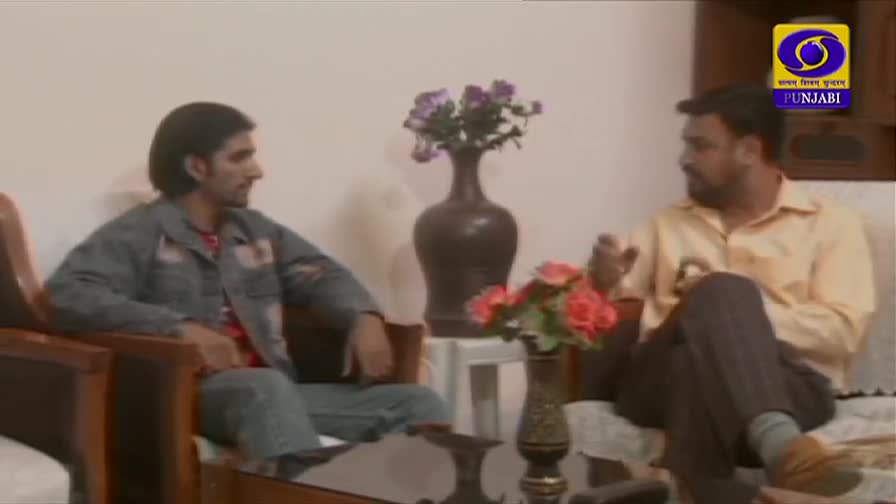 Two men sit in a living room, engaged in conversation, with a DD Punjabi logo visible in the corner. One gestures with his hands as he speaks, while the other listens intently.
