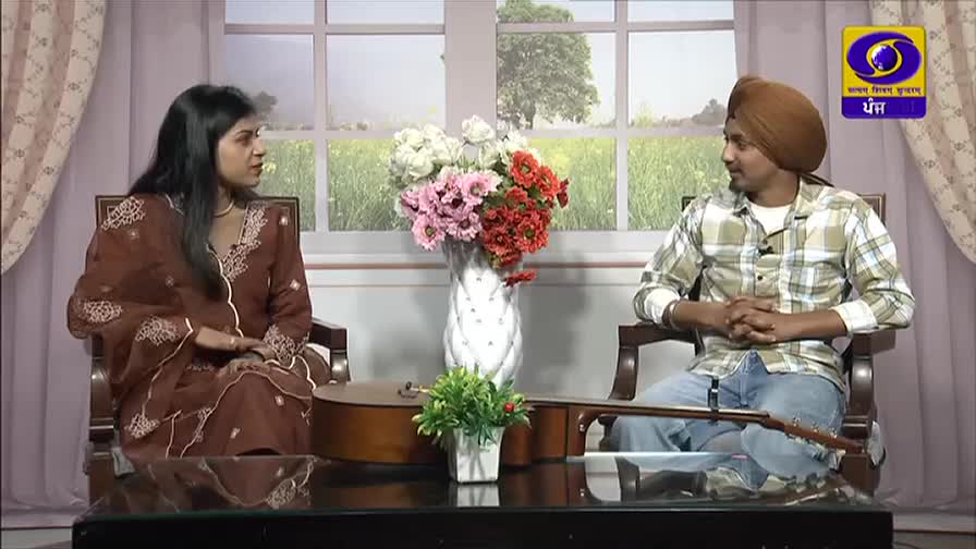 A woman in a brown salwar kameez and a man in a turban are seated in chairs, facing each other. A guitar rests on the table between them, and a logo for DD Punjabi is visible in the corner.
