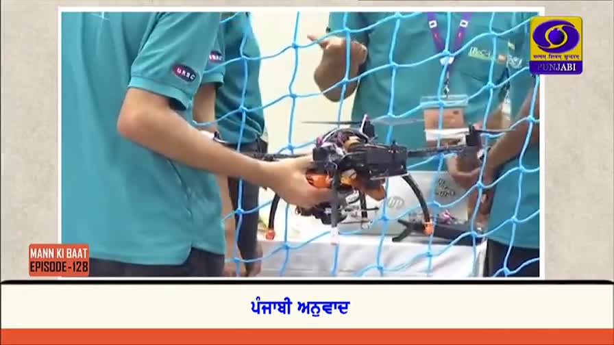 A person in a teal shirt carefully holds a small, black drone, its orange arms and wires visible. The scene is framed by a blue net, and the DD Punjabi logo is in the corner.
