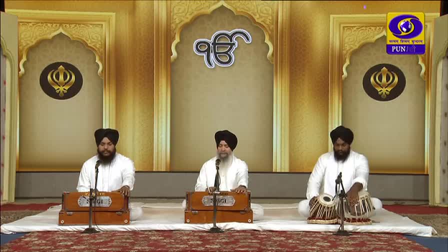 Three men in white robes and turbans sit on a platform, each with a microphone in front of them. The center man is singing into his microphone, while the other two play instruments.
